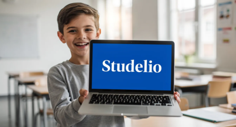 Un collégien souriant tient son ordinateur portable, sur lequel est inscrit "Studelio" en blanc sur un fond bleu vif. Le jeune élève exprime sa joie et son enthousiasme pour ses cours et ses exercices de français.